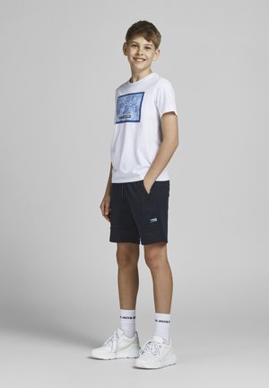 Camiseta blanca con diseño gráfico azul, pantalones cortos azul marino con bolsillos, zapatillas blancas y calcetines blancos con marca negra. Fondo gris liso.