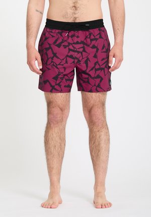 Homme portant un short de bain bordeaux mi-cuisse avec un motif abstrait noir et une ceinture noire, debout pieds nus sur un fond uni.