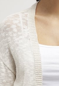 Lekki kardigan dziany w kremowym kolorze, z prążkowanym dekoltem, luźnym krojem i teksturowanym wzorem. Nałożony na białą koszulkę bez rękawów.