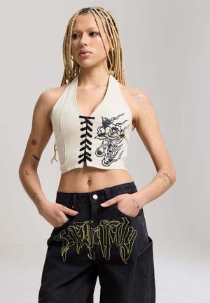 Junge Person mit langen blonden Zöpfen, trägt ein ärmelloses weißes Crop-Top mit schwarzem Blumenmuster und schwarzen Hosen mit gelber Beschriftung.