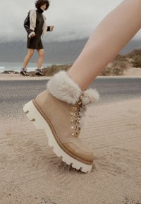 Botas de tobillo marrones con un puño de felpa beige, ojales de metal y suela gruesa color crema. Material de ante con un detalle de ribete cosido.