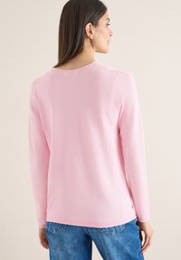 Femme aux longs cheveux bruns portant un haut à manches longues texturé rose clair et un jean bleu, vue de dos sur un fond beige.