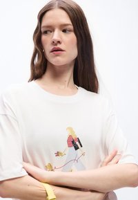 Γυναίκα με μακριά καστανά μαλλιά, φοράει λευκό μπλουζάκι με γραφικό μιας ξανθιάς γυναίκας που περπατάει ένα σκύλο, με σταυρωμένα τα χέρια, κοιτάζει προς το πλάι.