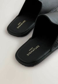 Black slide-on slippers made from textured leather, featuring a smooth footbed and gold branding "NEXT SIGNATURE" on the insole.