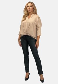 Beżowa, luźna bluzka z kołnierzykiem i fałdą z przodu, połączona z czarnymi, dopasowanymi spodniami z ekoskóry i czarnymi wysokimi obcasami.