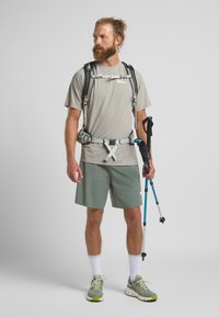 T-shirt léger gris clair évacuant l'humidité avec un col rond, associé à un short vert. Équipé d'un sac à dos de randonnée et d'un bâton de trekking.