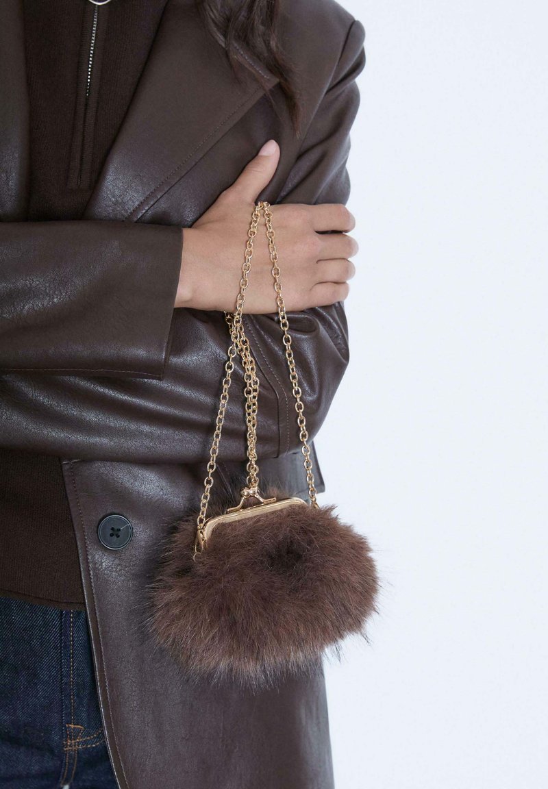 Borsa in pelliccia sintetica marrone con tracolla in catena dorata e chiusura a morsetto. La borsa è rotonda e morbida, appoggiata su una giacca di pelle scura.