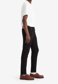 Vaqueros negros ajustados, camiseta blanca y zapatos marrones con cordones. Detalles notables incluyen un cinturón beige y costuras visibles en los vaqueros.