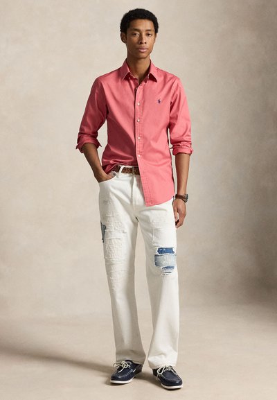 Νέος άνδρας με σγουρά μαλλιά στέκεται φορώντας κοραλλί πουκάμισο με γυρισμένα μανίκια, λευκά τζιν με μπαλώματα, καφέ ζώνη, ρολόι και ναυτικά παπούτσια σε σκούρο μπλε.