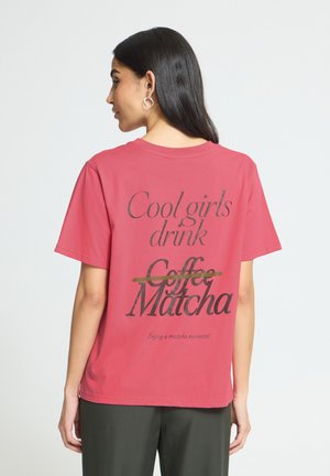Femme aux longs cheveux foncés portant un T-shirt rouge avec le texte "Cool girls drink Matcha," où "Coffee" est barré, et un pantalon noir, de profil.