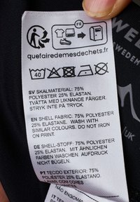 Clothing care label, white fabric with black text, detailing material composition, washing instructions, and care symbols. Black background.