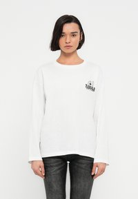 Γυναίκα με κοντά σκούρα μαλλιά, που φοράει λευκό πουκάμισο με μακριά μανίκια με το κείμενο και το γραφικό "Bagels & Coffee" στο στήθος, σε συνδυασμό με μαύρο παντελόνι.