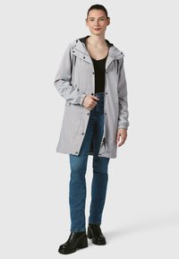 Lys grå vandtæt jakke med snøring i hætten, lynlås lukning og elastiske manchetter, sammen med mørkeblå jeans og sorte højhælede støvler.
