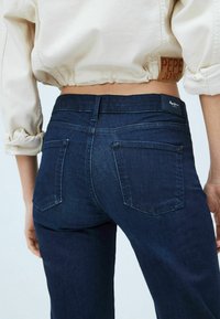 Tummansinisiä farkkuja, joissa on keskipitkä vyötärö, kaksi takataskua ja pieni musta etiketti vyötäröllä, kuvattuna takaa.