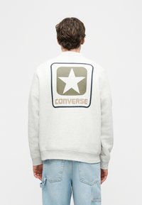 Άνδρας με πλάτη, φοράει γκρι ανοιχτό φούτερ με μεγάλο λογότυπο του αστεριού Converse σε ελιά και μπεζ χρώμα στην πλάτη και ανοιχτό μπλε τζιν παντελόνι.