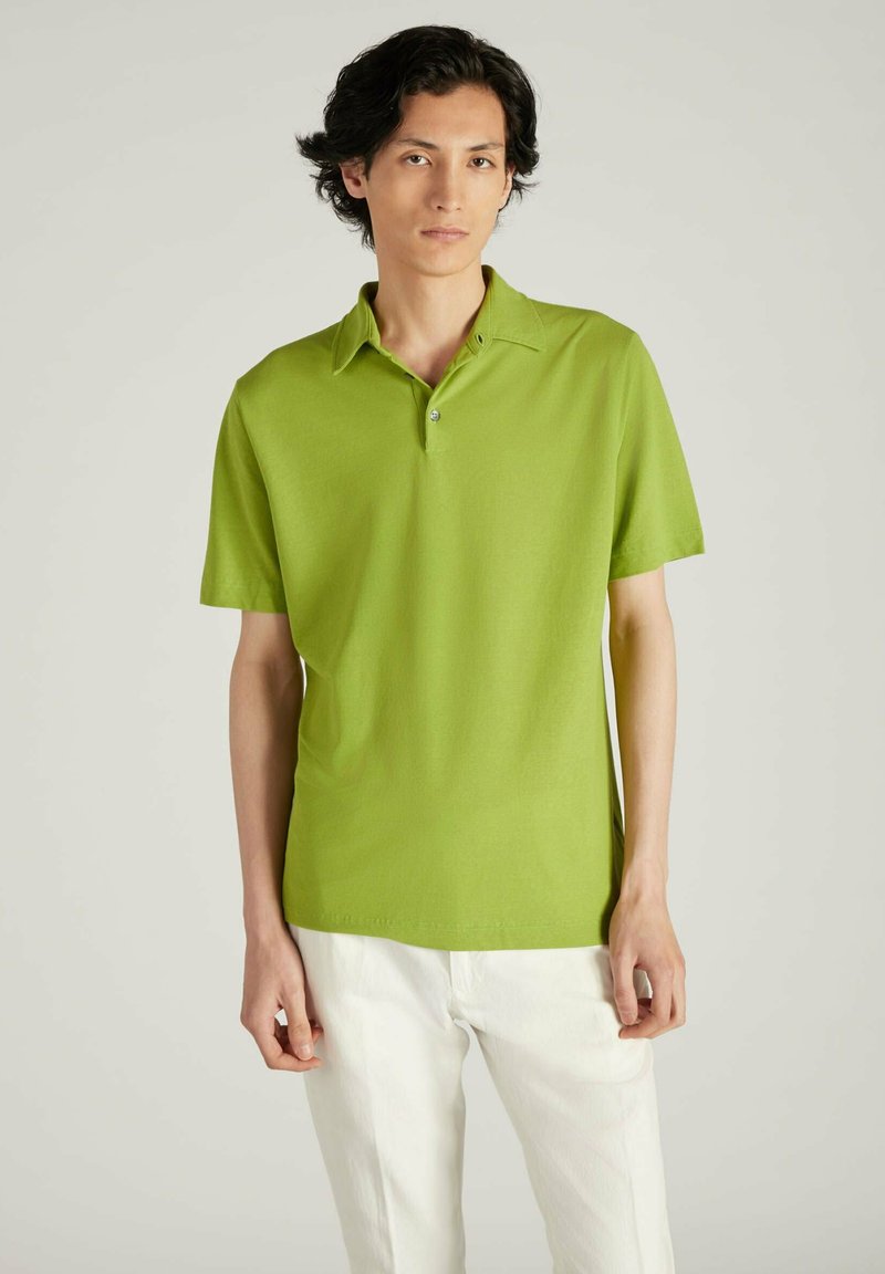 Polo de algodón verde con mangas cortas y una tapeta de dos botones. Se lleva con pantalones blancos, presenta líneas limpias y un detallado minimalista.