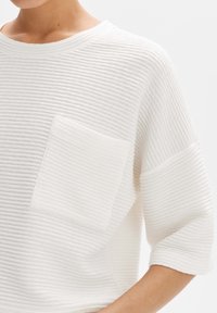 Top de punto blanco con canalé, cuello redondo, mangas cortas y un bolsillo de parche. Presenta una tela texturizada y patrones de canalé horizontales.