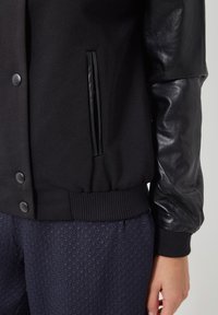 Chaqueta bomber negra con tejido texturizado, mangas de cuero liso, cierre de botones a presión y dobladillo y puños acanalados.