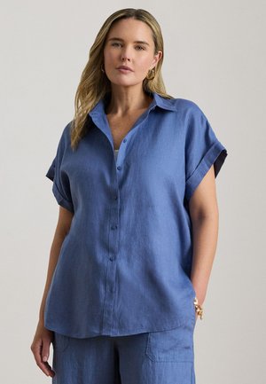 Zila linu pogāžamā krekla ar īsām sarullētām piedurknēm, ar vaļēju piegriezumu un retu pogu rindu priekšpusē. Gluda tekstūra, minimālistisks dizains.