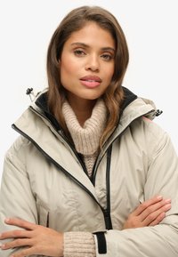 Beige geïsoleerde jas met een hoge kraag, zwarte rits en zwarte accenten. Zacht crème trui eronder. Model heeft de handen gekruist.