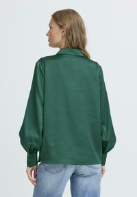 Blusa di raso verde smeraldo con maniche lunghe a sbuffo, colletto e polsini abbottonati. Texture liscia, vestibilità leggermente ampia, abbinata a jeans azzurri chiari.