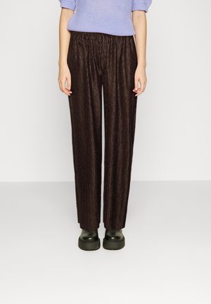 Vrouw draagt losse, donkerbruine, gestructureerde broek, zwarte grove laarzen en een lichtpaars gebreid topje, staand tegen een effen lichte achtergrond.