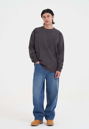 Grijze long-sleeve shirt met grafische ontwerpen, gecombineerd met losse blauwe jeans en bruine klompen, die een ontspannen pasvorm en casual stijl benadrukken.