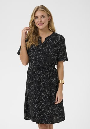 Woman with long blonde hair wears black short-sleeve dress with white polka dots, accessorized with gold bracelet, rings, and hoop earrings.
