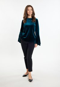 Top lungo a maniche con polsini svasati in velluto verde acqua, abbinato a pantaloni neri slim-fit e tacchi a punta neri, su uno sfondo bianco.