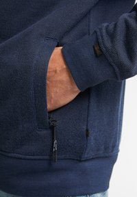 Pulover bleumarin cu un model texturat de tricot. Are o buzunar cu fermoar pe lateral și un manșon cu un model în relief. Eticheta handmade este vizibilă pe încheietura mâinii.