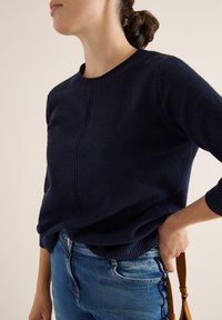 Dunkelblauer Pullover aus weichem Strick mit Rundhalsausschnitt, gerippten Bündchen und Nahtdetails an der Vorderseite. Kombiniert mit hellblauen Jeans.