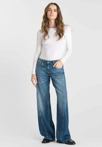 Haut blanc léger à manches longues associé à un jean en denim bleu moyen à jambes larges avec une finition délavée et un design à cinq poches. Chaussures plates noires.