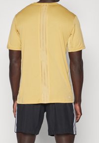 Muž oblečený v žlutém sportovním tričku s krátkým rukávem a vertikálními pruhy, a v černých šortkách s bílými bočními pruhy, pohledem zezadu.