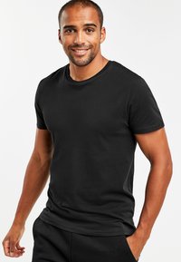 Tricou negru cu mânecă scurtă, dintr-un material moale, cu guler rotund, siluetă ajustată și textură netedă, purtat într-un stil casual.