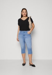Svart kortermet topp med bølget V-hals, lyseblå avskårne jeans og svarte slip-on sko. Enkel design, glatt tekstur og tettsittende silhuett.