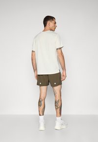 Light-colored short-sleeve t-shirt and dark green shorts with white circular accents; tattoos visible on legs; white sneakers.