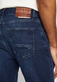 Tmavě modré džíny z denimu mají texturovanou hnědou koženou nášivku na pase, s malým detailem loga v červené, bílé a modré barvě poblíž zadní kapsy.