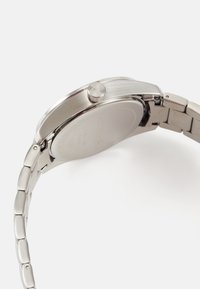 Ceas din oțel inoxidabil argintiu, cu carcasă rotundă, bezel lucios, coroana texturată și brățară din linkuri. Spate transparent al carcasei cu detaliu de logo.
