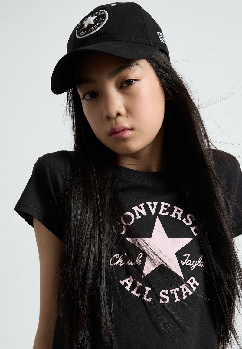 Sort t-shirt med et pink stjerne grafik og teksten "Converse All Star". En sort kasket med et rundt logo fuldender looket.