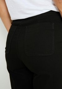 Vaqueros negros con una textura de tela suave, corte ajustado y dos bolsillos traseros con detalles de costura en contraste.