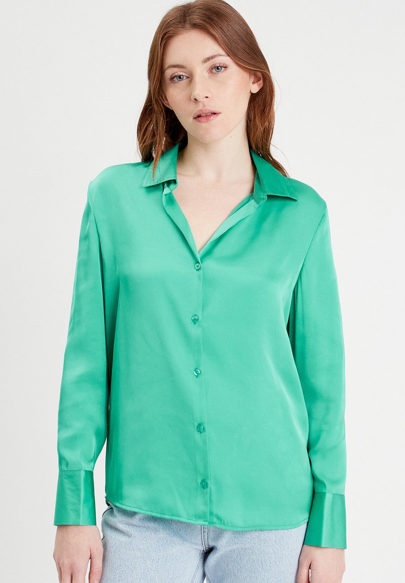 Chemise en satin vert avec col, manches longues et texture lisse. Présente un ourlet légèrement courbé et des boutons assortis.