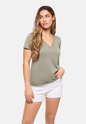 Mujer con cabello largo y ondulado que lleva una camiseta de cuello en V verde oliva y pantalones cortos blancos remangados, de pie con una mano en el bolsillo.
