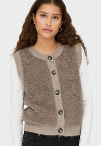 Flauschige braune ärmellose Strickjacke mit geripptem Saum, ausgestattet mit fünf dunkelgrünen Knöpfen auf der Vorderseite. Getragen über einem weißen Langarmshirt.