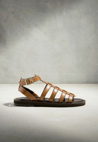 Sandale gladiateur en cuir marron avec bords festonnés et boucle argentée sur une semelle plate noire sur fond neutre.