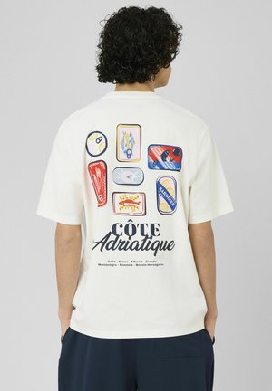 Personne aux cheveux bouclés portant un t-shirt blanc avec des illustrations colorées de poissons en conserve et le texte "Côte Adriatique" dans le dos.