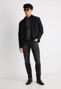 Νέος άντρας που φοράει σκούρα γυαλιά ηλίου, μαύρο σακάκι, ξεθωριασμένο μαύρο τζιν, μαύρη ζώνη και μαύρα μποτάκια αστραγάλου, στέκεται με το ένα χέρι στην τσέπη.