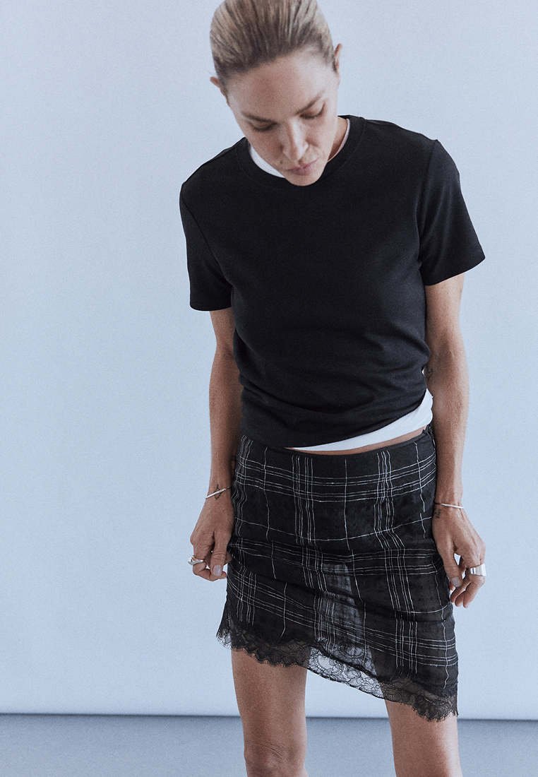 Femme aux cheveux clairs attachés portant un t-shirt noir à manches courtes et une jupe noire à carreaux avec une bordure en dentelle, debout devant un fond uni.