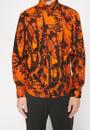 Camisa de manga larga con botones, con estampado de hojas y ramas negras y rojas sobre fondo naranja brillante, combinada con pantalones negros.