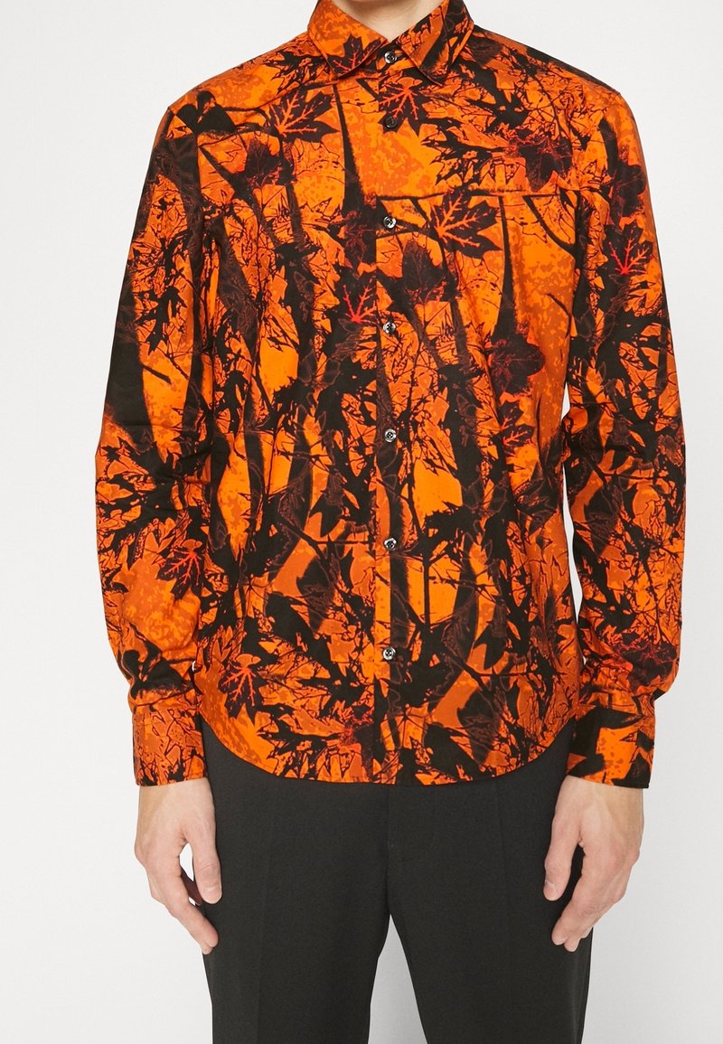 Chemise à manches longues boutonnée avec motif de feuilles et branches noires et rouges sur fond orange vif, portée avec un pantalon noir.