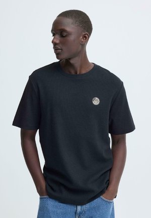 Zwarte korte mouw T-shirt met een getextureerde stof, ronde hals en een klein cirkelvormig logo op de borst. Gecombineerd met blauwe spijkerbroek.